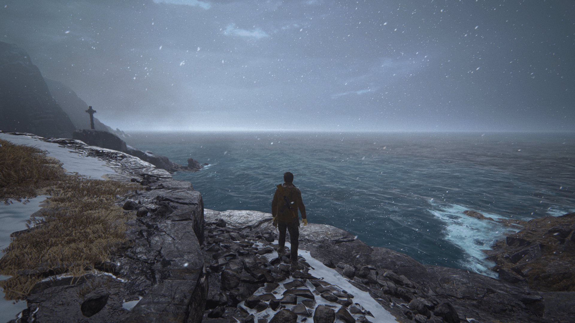 A man stands on a rocky, snow-dusted cliff edge overlooking a stormy ocean, with snow falling and a large stone cross visible in the distance.