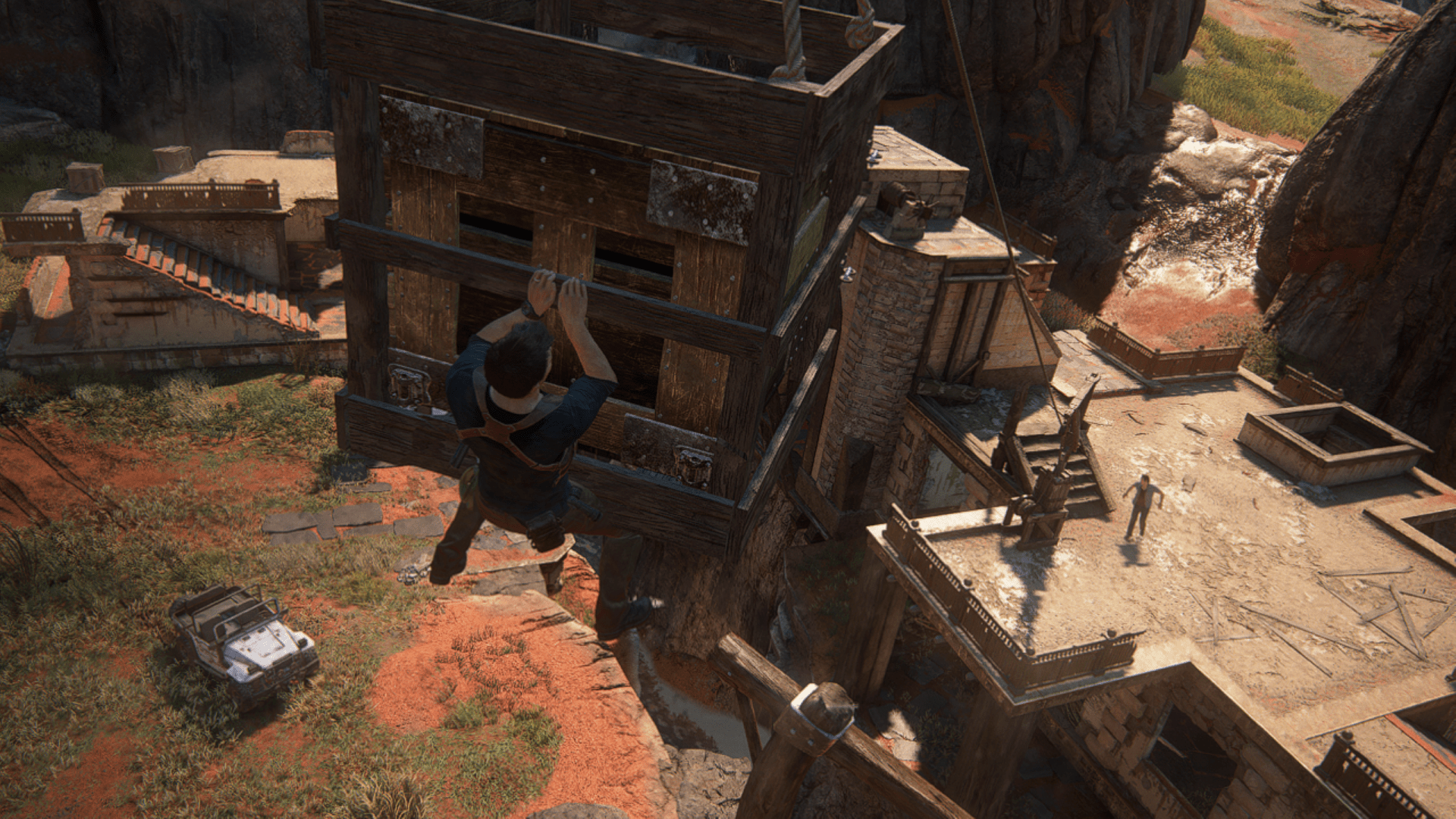 A man clings to a suspended wooden crate above a ruin, while another figure stands on a rooftop below.